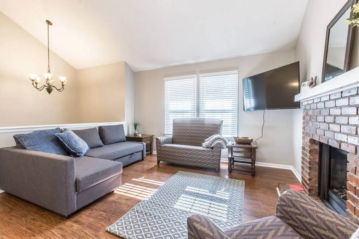 A cozy living room scene centers on a plush sofa and armchair facing a fireplace and television, accented by a stylish chandelier. The arrangement is ideal for relaxing, entertaining friends, or enjoying a movie night with family. The overall look is contemporary, with thoughtful decor touches throughout.