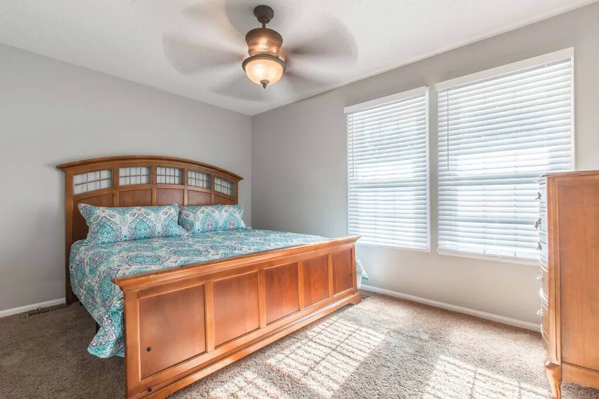 A serene bedroom featuring a wooden bed frame with colorful bedding, illuminated by natural light streaming in through large windows. The space feels quiet, restful, and well-suited for relaxation or a deep night's sleep.