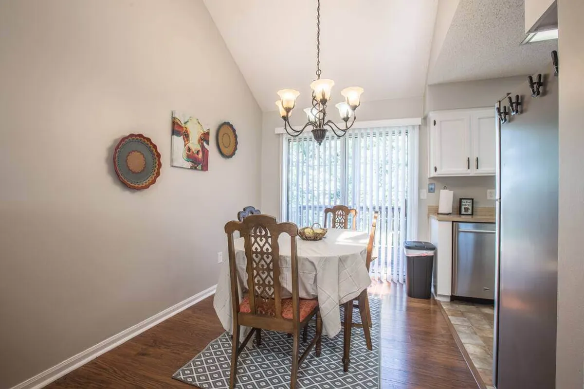 A warm dining area with a large table, an elegant chandelier, colorful wall art, and a sliding glass door that opens to the outdoors. The space is set for lively meals and shared moments, with plenty of natural light inviting the outside in.