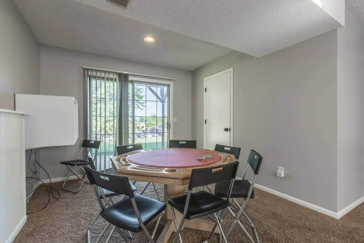 A bright, casual space featuring a round poker table surrounded by six black chairs, set near large windows that flood the room with natural light. The open feel and inviting setup suggest it's ideal for group fun and laughter.
