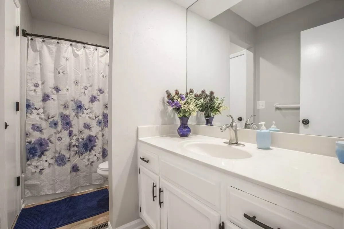 This image showcases a modern bathroom with a sleek vanity and sink, complemented by a decorative vase with fresh flowers and a clean shower curtain in the background. The abundance of natural light and uncluttered surfaces emphasize the fresh finishes and calm, inviting atmosphere.