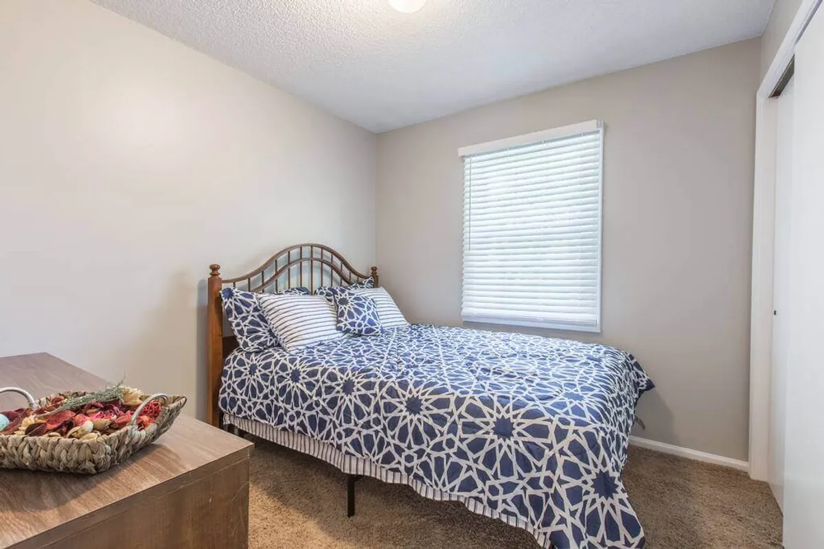 A tranquil bedroom with a bed featuring patterned bedding, a window with adjustable blinds that lets in gentle daylight, and a nightstand with a decorative basket. The peaceful environment is ideal for uninterrupted rest or focused, heads-down work.