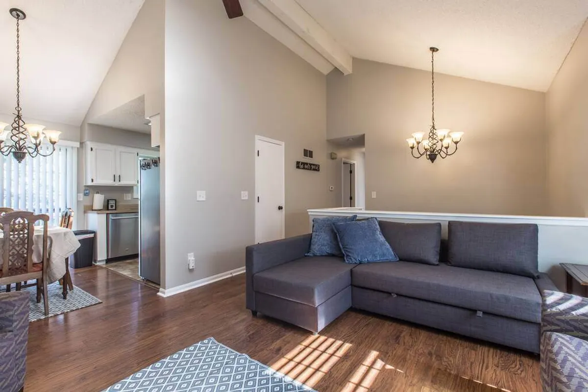 This image presents a comfortable sectional sofa beneath a modern chandelier, with a view extending to a spacious dining table. The open layout seamlessly connects lounging and dining spaces, capturing the essence of open-concept living. Warm lighting and tasteful decor create an inviting setting for both small gatherings and lively celebrations.
